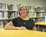 Woman smiling in a library, wearing a pink shirt. Bookshelves and blurred signs are visible in the background. Warm, inviting atmosphere.