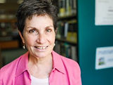 Woman smiling in a library, wearing a pink shirt. Bookshelves and blurred signs are visible in the background. Warm, inviting atmosphere.